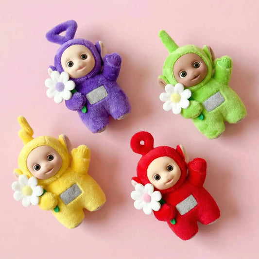 Ensemble de quatre charms Teletubbies en peluche (Tinky Winky, Dipsy, Laa-Laa, Po), chacun tenant une petite fleur blanche, disposés en losange sur un fond rose uni.