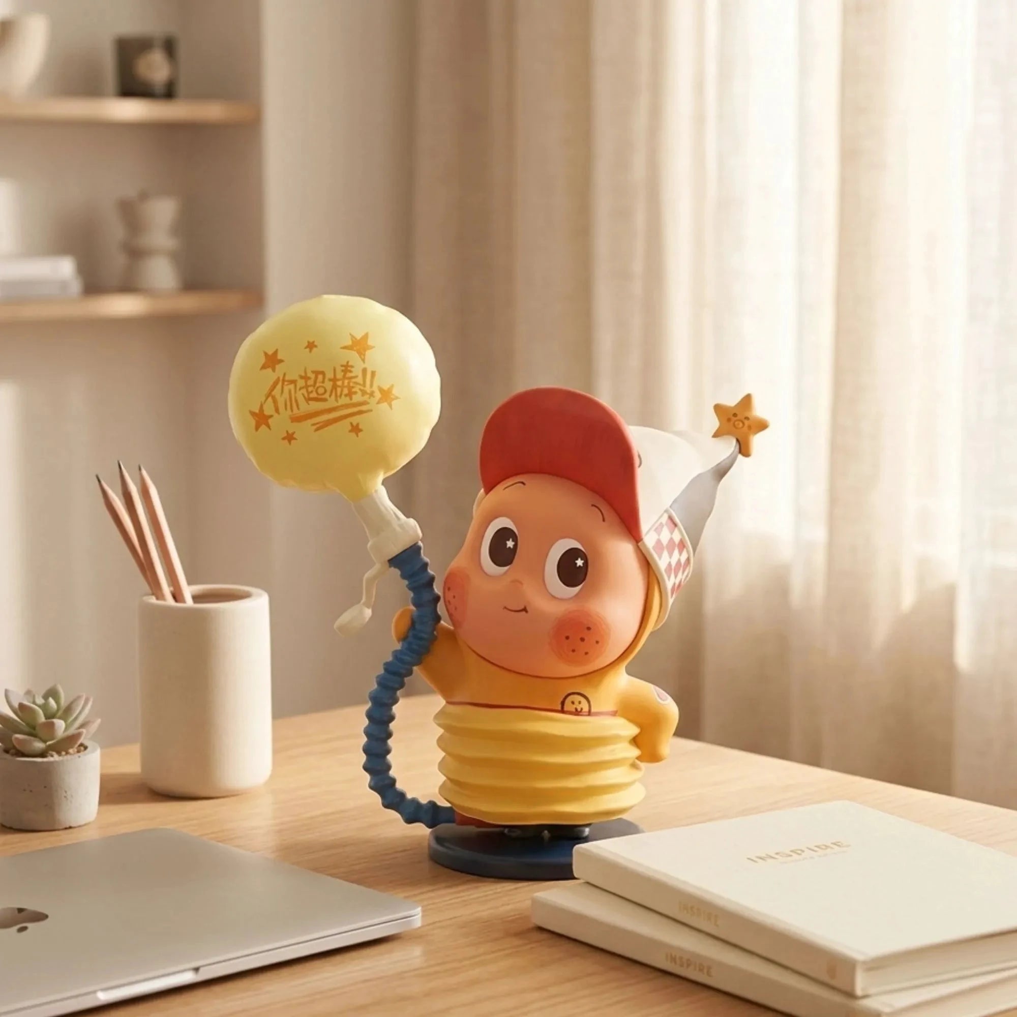 La figurine 'Little Balloon' de Star Kid est posée au centre d'un bureau en bois d'inspiration japonaise-scandinave. L'atmosphère est chaleureuse et lumineuse grâce à la lumière naturelle venant d'une fenêtre à droite. À gauche de la figurine, on voit un pot à crayons en céramique blanche et une petite plante grasse en pot. Au premier plan, le coin d'un ordinateur portable gris et deux cahiers 'INSPIRE' blancs sont empilés.