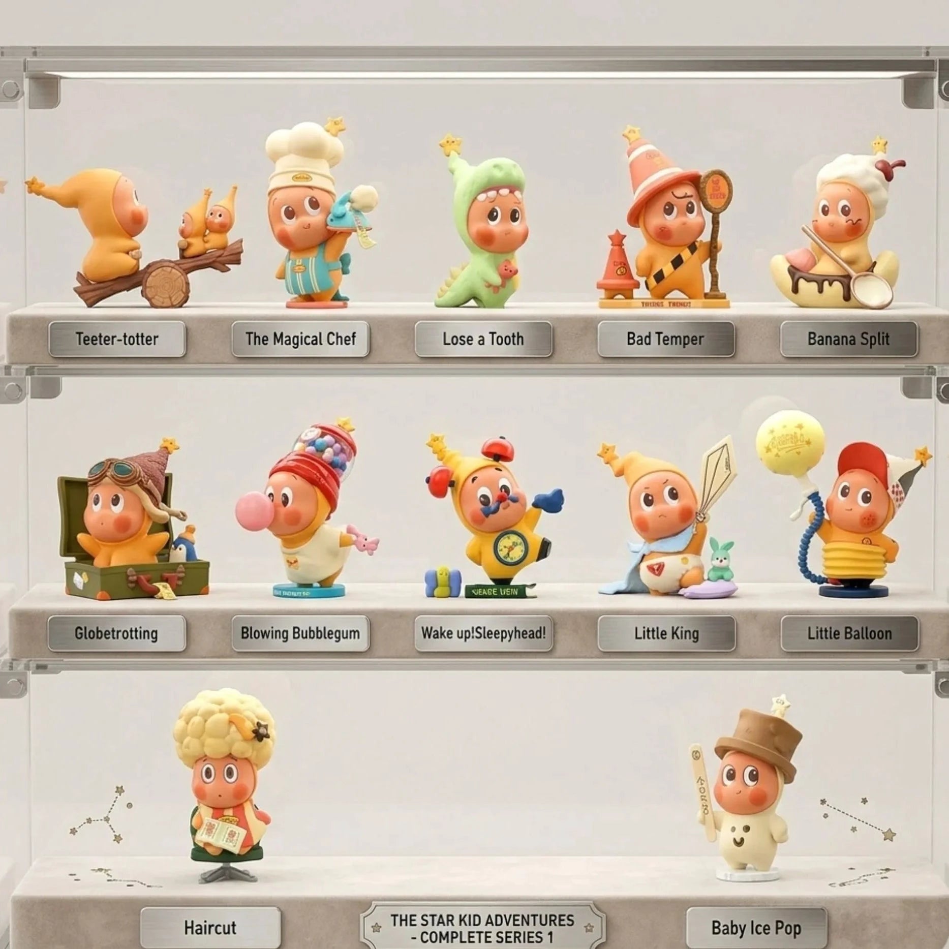 Une vitrine de collection transparente en acrylique à trois étagères contenant la collection 'The Star Kid Adventures - Complete Series 1'. Chaque étagère présente des figurines en vinyle distinctes avec des étiquettes individuelles en métal comme 'Teeter-totter', 'Globetrotting', et 'Little King'. L'étagère du milieu contient la figurine 'Little Balloon'. Les étagères sont éclairées par des lumières LED douces et intégrées. L'arrière-plan de la vitrine présente un motif de constellation subtil.